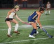 Field hockey players in action during a night game on grass, one in white, one in blue, competing for the ball.