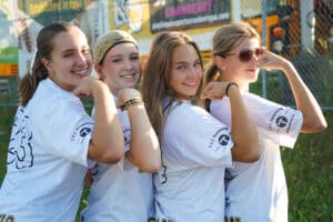 Four smiling girls in matching t-shirts posing with flexed arms outdoors.