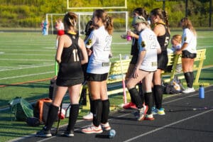Field hockey players take a break on the sidelines during a game on a sunny day.