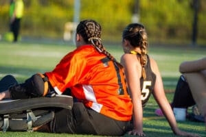 Two female field hockey players with braided hair sitting on the sidelines during a break.