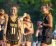 Teen field hockey players in black Tigers jerseys, smiling before a game, bright sunlight, green trees in background.