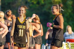 Teen field hockey players in black Tigers jerseys, smiling before a game, bright sunlight, green trees in background.