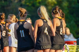 Girls in black sports jerseys wait on the field, ready for a team activity in the sunshine.