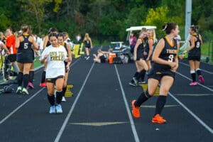 Girls warming up for field hockey practice on a track, wearing sports uniforms and gear.