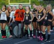 Field hockey team posing on sidelines, players in uniforms holding sticks, happy expressions, ready for the game.