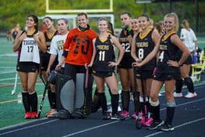 Field hockey team posing on sidelines, players in uniforms holding sticks, happy expressions, ready for the game.
