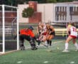 Field hockey match with goalie blocking a shot, players in action, and ball near the goal.