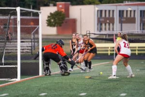 Field hockey match with goalie blocking a shot, players in action, and ball near the goal.