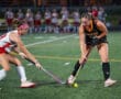 Two field hockey players compete for the ball during a match on a green turf field.