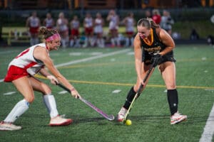Two field hockey players compete for the ball during a match on a green turf field.