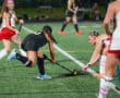 Field hockey players compete intensely for the ball on a bright green turf under stadium lights.