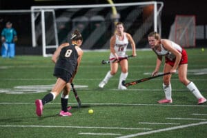 Field hockey match action with player in black dribbling past opponents in white on a vibrant green turf.