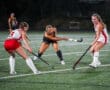 Girls playing field hockey on turf, focused on a black dress player making a pass. Sports action shot at night game.