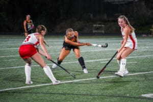 Girls playing field hockey on turf, focused on a black dress player making a pass. Sports action shot at night game.