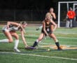 Two field hockey players compete for the ball on a turf field during a night match.