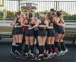 Girls' field hockey team huddling in circle, showing unity and motivation before a game on the stadium field.
