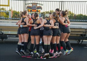 Girls' field hockey team huddling in circle, showing unity and motivation before a game on the stadium field.