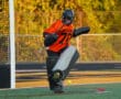 Field hockey goalie in action, blocking a ball during a sunny match, wearing orange and black protective gear.