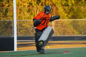 Field hockey goalie in action, blocking a ball during a sunny match, wearing orange and black protective gear.