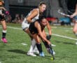 Female field hockey players competing during a match on a turf field at night.