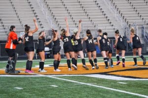 Field hockey team celebrates on the field, raising arms, showing unity and sportsmanship before a game.