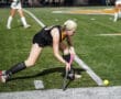 Field hockey player in action, wearing black uniform, bending to hit a yellow ball on a green turf field.