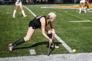Field hockey player in action, wearing black uniform, bending to hit a yellow ball on a green turf field.