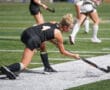 Field hockey player in black uniform striking the ball on artificial turf during a match.