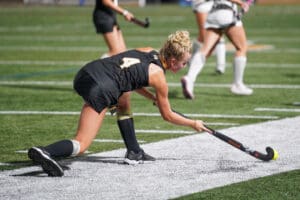 Field hockey player in black uniform striking the ball on artificial turf during a match.