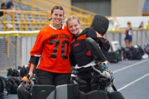 Two field hockey goalkeepers in black and orange gear smiling together on the sideline.