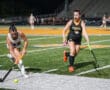 Two field hockey players in action during a night game on a turf field.