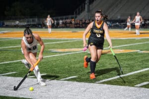 Two field hockey players in action during a night game on a turf field.