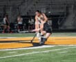 Two female field hockey players compete for the ball on a brightly lit stadium field.