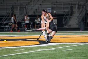 Two female field hockey players compete for the ball on a brightly lit stadium field.
