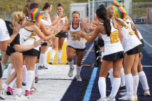 Field hockey player wearing jersey 34 runs through cheering tunnel, surrounded by teammates in colorful hats.
