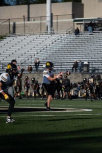 Football players catching a pass during a game on a sunny day, with empty bleachers in the background.