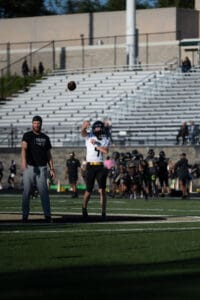 Football player in action throws a pass on the field during practice, with coach observing.