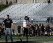 Football player warming up on the field with coach nearby; empty bleachers in the background.