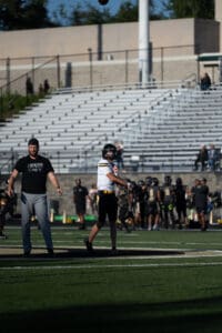 Football player warming up on the field with coach nearby; empty bleachers in the background.