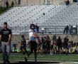 Football player practicing throw on field with coach observing, empty bleachers in background. Football game preparation.