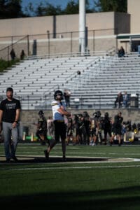Football player practicing throw on field with coach observing, empty bleachers in background. Football game preparation.