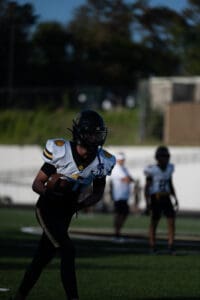 Football player running on field with ball during a game, wearing black and white uniform, helmet, daytime practice.