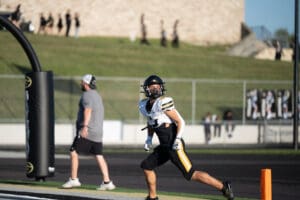 Football player in action near end zone during a game, with a coach in the background on a sunny day.