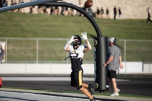 Football player catching a pass in the end zone during a game, with a coach observing from the sidelines.