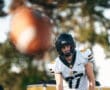 American football player in a helmet observing a moving football during practice outdoors.