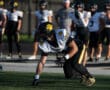 Football player crouching on field, preparing for a play during practice. Teammates in background.