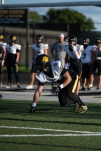Football player crouching on field, preparing for a play during practice. Teammates in background.