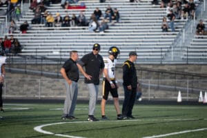 Football coaches and player on the sideline during a game with spectators in background.