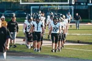 Football team in black uniforms entering field, lined up for game preparation, sunny day.