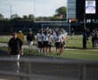 Football team walking onto field under clear sky, nearing scoreboard, in black and white uniforms.
